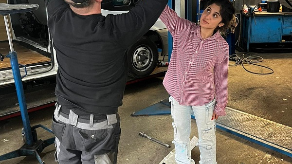 Eine Person arbeitet unter einem Fahrzeug und benutzt ein Werkzeug, während eine andere Person zuschaut und das Geschehen aufmerksam verfolgt. Die Szenerie befindet sich in einer Werkstatt mit mehreren Werkzeugen und Geräten im Hintergrund.