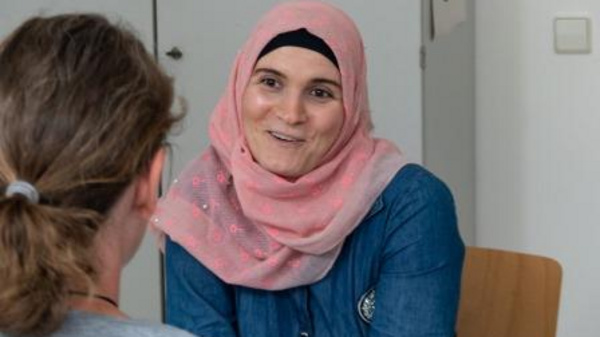 A woman wearing a pink hijab and a denim top smiles while engaging in conversation with another person. The setting suggests a friendly discussion, hinting at a supportive atmosphere.