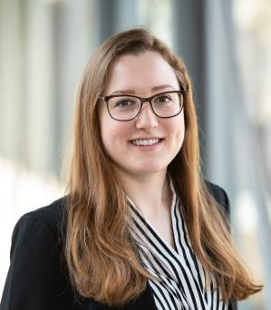 Eine junge Frau mit langen, hellbraunen Haaren und einer Brille lächelt in die Kamera. Sie trägt eine gestreifte Bluse und einen dunklen Blazer. Der Hintergrund zeigt eine helle, moderne Umgebung mit Fensterlicht.