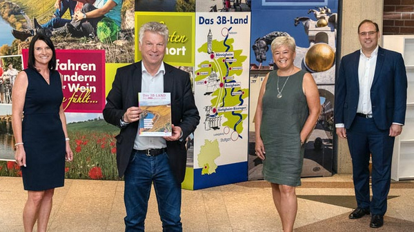 A group of five people stands in front of a backdrop featuring tourist information about the 3B region. The central figure holds a brochure while smiling. The others include a woman in a black dress, a woman with short hair, and two men, one wearing a casual outfit and the other in a suit.