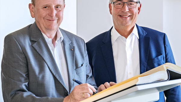 Zwei Männer stehen nebeneinander, lächelnd in einem hellen Raum. Der Mann links trägt einen grauen Anzug und hält einen Stift, während der Mann rechts ein weißes Hemd und einen blauen Blazer trägt. Auf einem Tisch zwischen ihnen liegt ein aufgeschlagenes Buch.