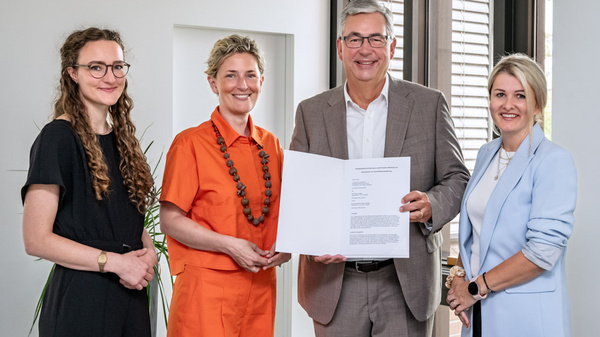 Vier Personen posieren in einem modernen Raum. Eine Frau in orangefarbener Kleidung hält ein Dokument in der Hand, während die anderen drei Personen, in formeller Kleidung, freundlich lächeln und zusehen. Ihre Gesichtsausdrücke vermitteln einen positiven und feierlichen Moment.