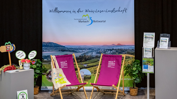Ein Empfangsbereich mit einem blauen Hintergrund, auf dem „Willkommen in der Weserbergland“ steht. Vorne stehen zwei bequeme Liegestühle. Links und rechts sind Informationsstände mit Broschüren und Veranstaltungsflyern zu sehen, die über die Region informieren.