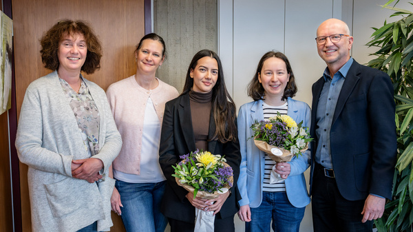 Fünf Personen stehen in einer Büroumgebung, lächeln und halten Blumensträuße in den Händen. Die Gruppe besteht aus vier Frauen und einem Mann, die in formeller Kleidung gekleidet sind. Der Mann steht rechts, die Frauen sind links angeordnet.