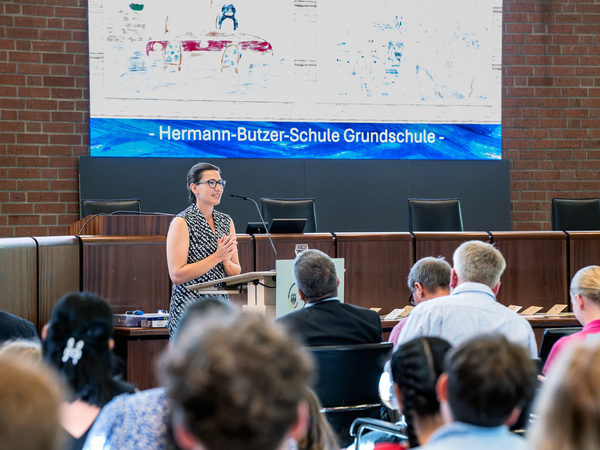 Eine Frau steht am Rednerpult und hält eine Ansprache in einem Saal mit Menschen, die ihr zuhören. Im Hintergrund ist ein Banner mit der Aufschrift "Hermann-Butzer-Schule Grundschule" zu sehen.