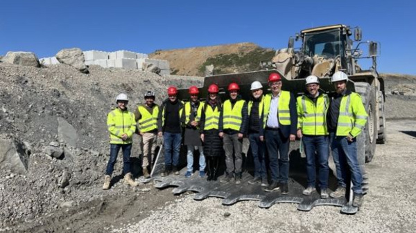 Eine Gruppe von zehn Personen steht auf einer Baustelle, umgeben von Schotter und einem großen Baumaschinenfahrzeug im Hintergrund. Alle tragen Warnwesten und Helm, einige auch Sicherheitsschuhe. Der Himmel ist blau und es handelt sich um eine sonnige Tagesaufnahme.