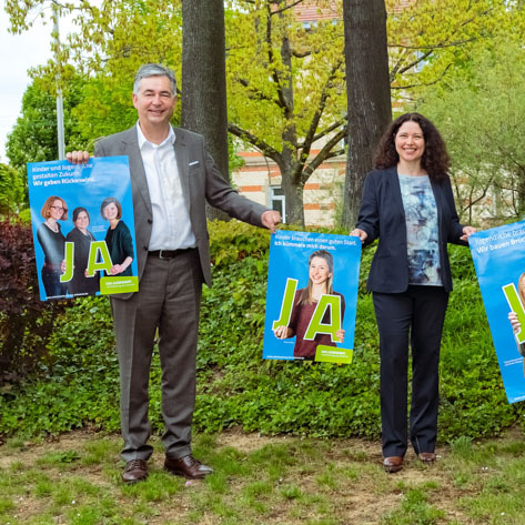 Vier Personen stehen draußen in einem Garten, jede hält ein Plakat mit dem Schriftzug „JA“. Sie lächeln und sind in formeller Kleidung. Im Hintergrund sind Bäume und Grünflächen zu sehen.