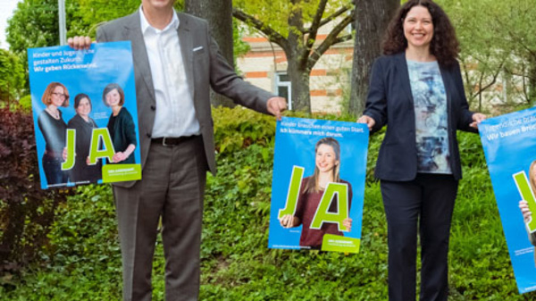 Four individuals stand outdoors, each holding a brightly colored poster featuring the word "JA!" in large letters. They appear engaged and supportive, with greenery and trees in the background, signifying a positive event or campaign.