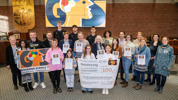 Eine Gruppe von Schülern und Lehrern steht in einem Raum und hält Zertifikate in den Händen. Im Vordergrund ist ein großes Banner mit dem Logo der „Gesunde Schule“ und ein Schild mit der Aufschrift „Belohnung 100€“ für die teilnehmenden Schulen.