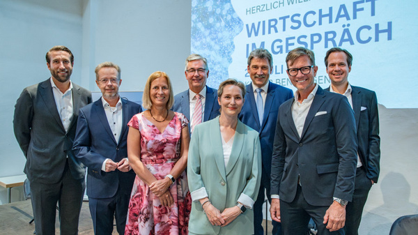 Eine Gruppe von acht Personen steht vor einem Präsentationshintergrund, auf dem „Wirtschaft im Gespräch“ steht. Die Menschen sind formell gekleidet und lächeln in die Kamera. Der Raum hat eine helle, freundliche Atmosphäre.