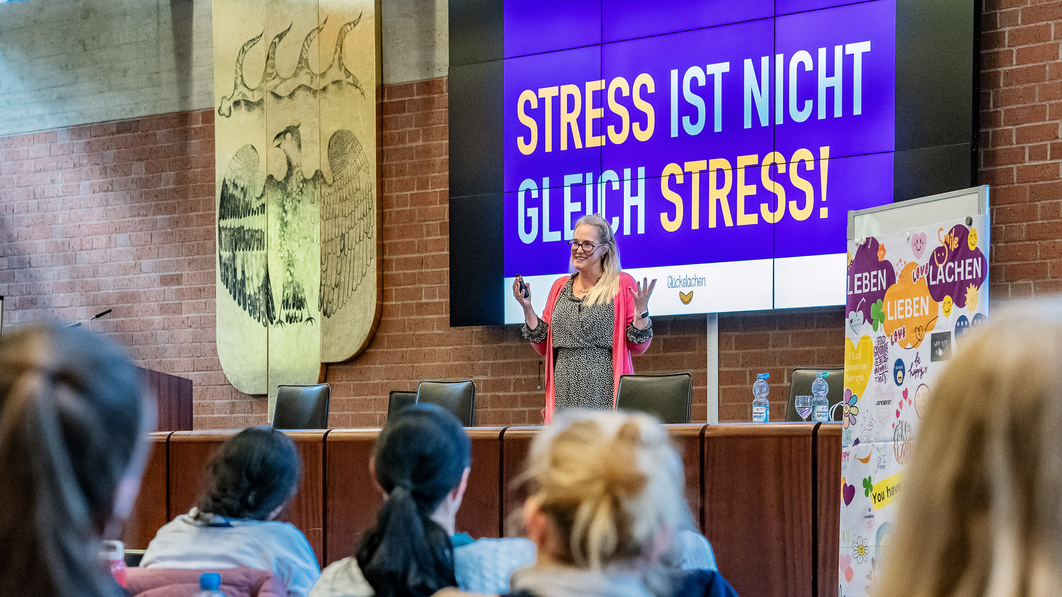 Speaker Nanni Glück stands behind a lectern and speaks with her hands raised; in the foreground you can see the backs of the heads of seated people.