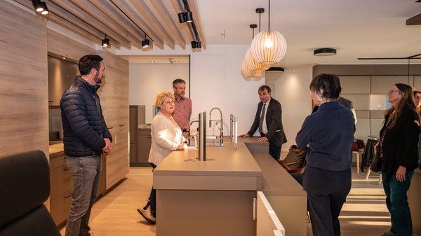 Eine Gruppe von fünf Personen steht um eine moderne Küchenarbeitsplatte. Sie diskutieren über die Ausstattung der Küche, die hellen Holz- und grauen Farbflächen sowie die modernen Lampen. Die Atmosphäre wirkt informativ und kooperativ.