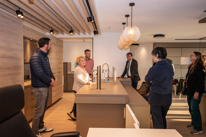 Eine Gruppe von fünf Personen steht um eine moderne Küchenarbeitsplatte. Sie diskutieren über die Ausstattung der Küche, die hellen Holz- und grauen Farbflächen sowie die modernen Lampen. Die Atmosphäre wirkt informativ und kooperativ.