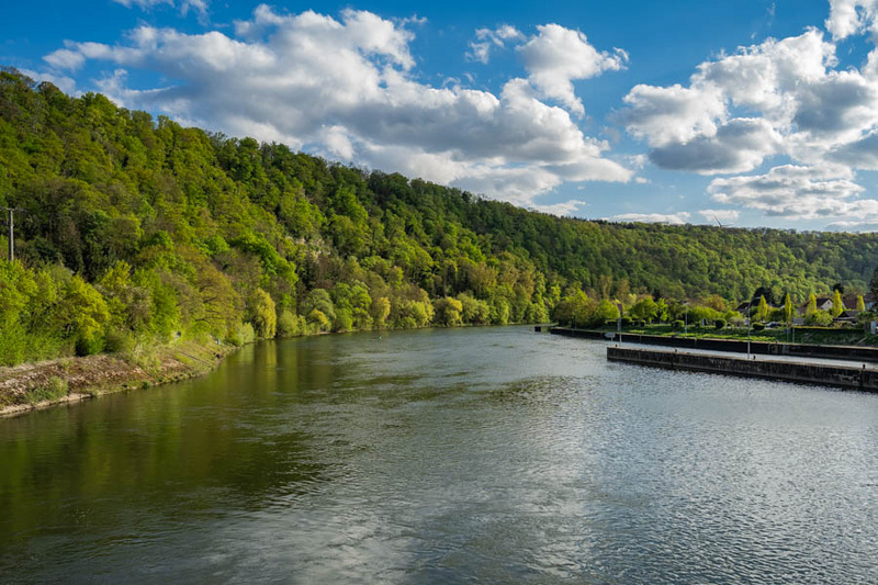 Ein ruhiger Fluss fließt durch eine grüne Landschaft, umgeben von bewaldeten Hügeln. Der Himmel ist teilweise bewölkt, mit vereinzelten Wolken, die über das Wasser reflektieren. Am Ufer gibt es einige Bäume und eine kleine Anlegestelle.