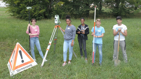 Five students stand in a grassy area, holding surveying equipment. They are positioned near a warning sign that reads "Vermessung." Each student appears engaged, showcasing a collaborative effort in a fieldwork activity related to land surveying.