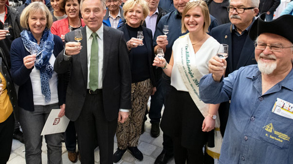 A group of diverse people stands together, smiling and holding glasses in a celebratory gesture. The scene suggests a festive atmosphere, possibly at a community event or gathering, with many individuals engaged and looking towards the camera.