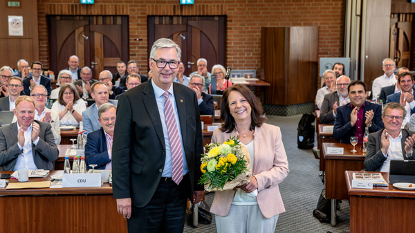 Eine Frau hält einen Blumenstrauß mit gelben Blumen, während ein Mann neben ihr steht. Im Hintergrund applaudiert eine Gruppe von Menschen in einem Konferenzraum. Die Atmosphäre ist feierlich und zeigt eine Anerkennung oder eine besondere Veranstaltung.
