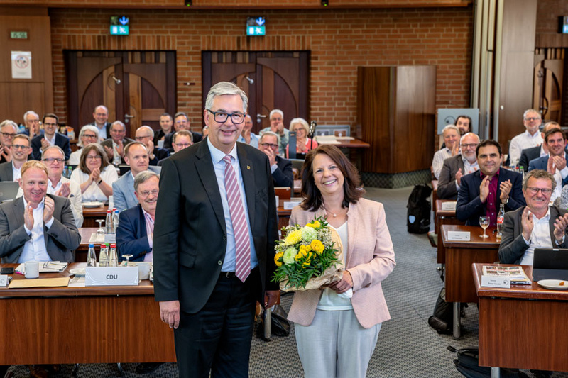 Eine Frau hält einen Blumenstrauß mit gelben Blumen, während ein Mann neben ihr steht. Im Hintergrund applaudiert eine Gruppe von Menschen in einem Konferenzraum. Die Atmosphäre ist feierlich und zeigt eine Anerkennung oder eine besondere Veranstaltung.