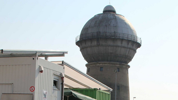 Ein großer Wasserturm mit einer runden, kuppelartigen Oberseite steht neben einem niedrigeren, modernen Gebäude. Der Wasserturm hat einen grauen Farbton und eine kleine Aussichtsplattform. Der Himmel ist klar und blau im Hintergrund.