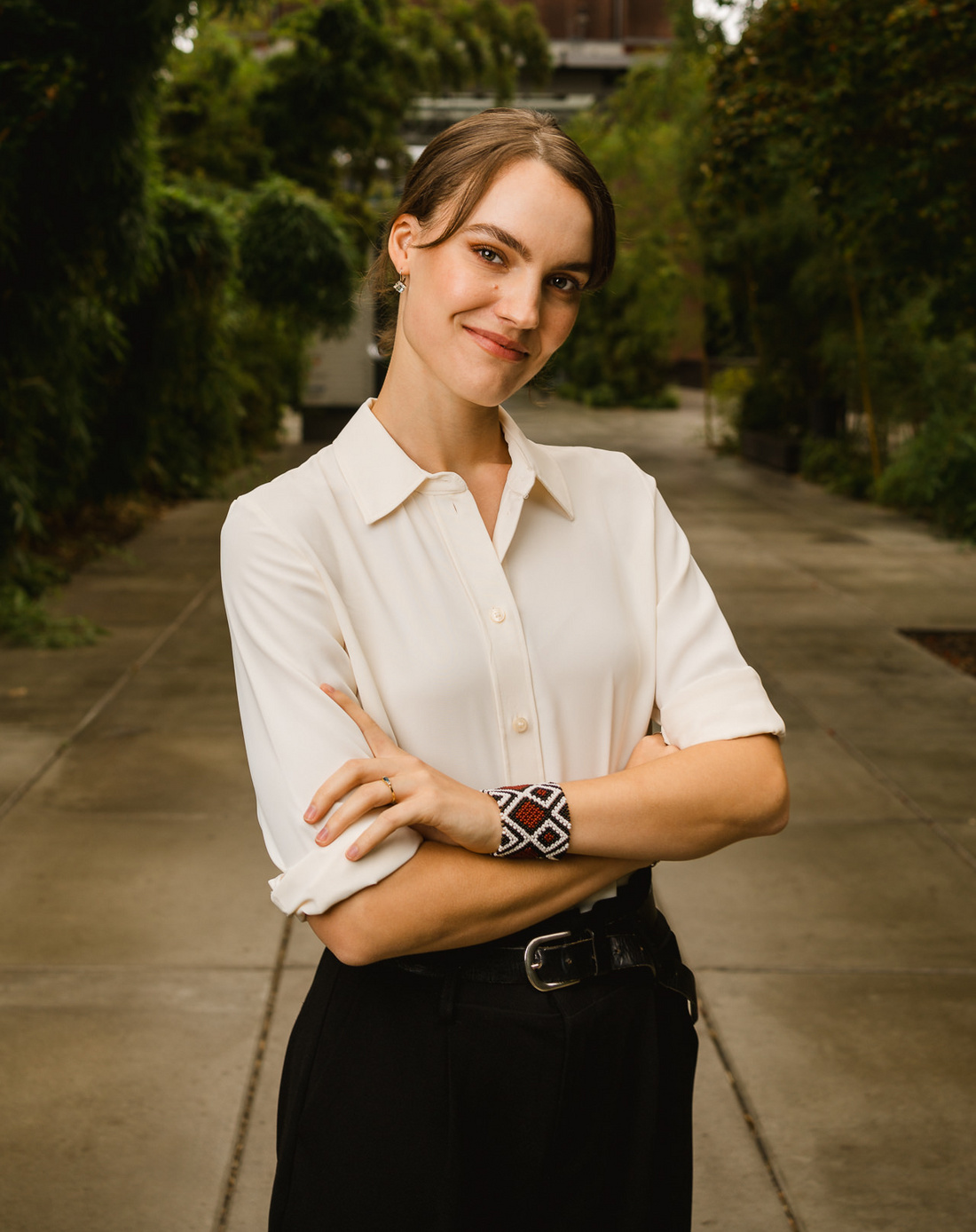 Eine Erwachsene Frau steht in einem Gartenbereich, lächelt und hat die Arme vor der Brust verschränkt. Sie trägt ein helles, kurzärmeliges Hemd und schwarze Hose. Im Hintergrund sind Grünpflanzen und ein moderner Gebäudeteil sichtbar.