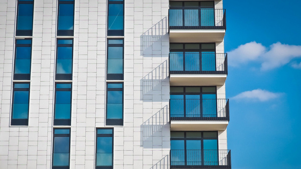 Ein modernes Wohngebäude mit einer hellen Fassade. Die rechte Seite zeigt mehrere Balkone mit dunklen Geländern und großen Fenstern, die einen Blick auf den Himmel ermöglichen. Wolken sind im Hintergrund sichtbar. (Text KI generiert)