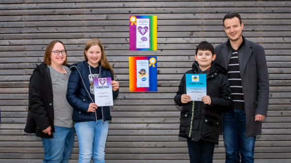 Sechs Personen stehen vor einer Holzwand und halten Urkunden oder Zertifikate in den Händen. In der Mitte der Gruppe sind mehrere Plakate oder Auszeichnungen aufgeklebt. Die Anwesenden lächeln und wirken stolz über ihre Erfolge oder Beiträge. (Text KI generiert)