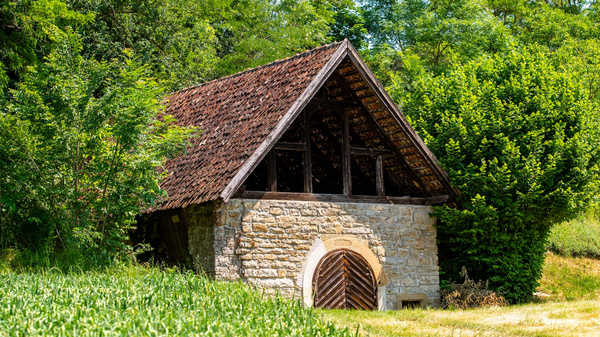 Ein einfaches, steinernes Gebäude mit einem dunklen, schrägen Dach steht inmitten von grünem Gras und Bäumen. Der Eingang ist mit einem großen, hölzernen Tor versehen. Die Umgebung wirkt ländlich und idyllisch.