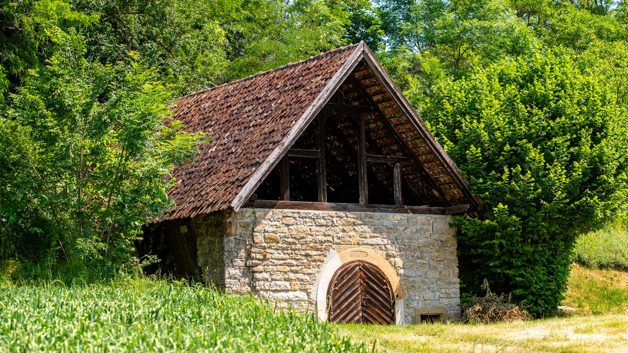 Ein einfaches, steinernes Gebäude mit einem dunklen, schrägen Dach steht inmitten von grünem Gras und Bäumen. Der Eingang ist mit einem großen, hölzernen Tor versehen. Die Umgebung wirkt ländlich und idyllisch.