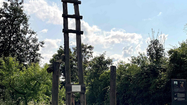 Ein hölzerner, schmaler Pfosten mit mehreren Sprossen steht auf einer Wiese. Umgeben von Bäumen und Sträuchern, erstreckt sich ein blauer Himmel mit vereinzelten Wolken darüber. Informationen zu dem Objekt sind an einem Schild neben dem Pfosten angebracht.