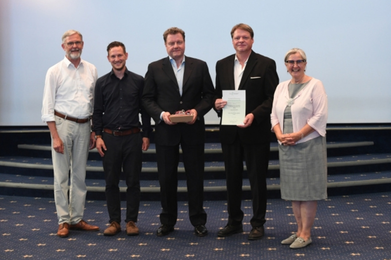 Fünf Personen stehen auf einer Bühne. In der Mitte hält ein Mann in einem schwarzen Anzug eine Urkunde und eine Auszeichnung. Die anderen vier Personen, in formeller Kleidung, sind um ihn herum positioniert und lächeln zur Kamera.