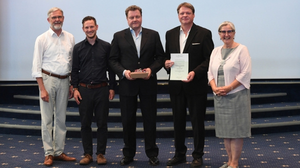 Fünf Personen stehen auf einer Bühne. In der Mitte hält ein Mann in einem schwarzen Anzug eine Urkunde und eine Auszeichnung. Die anderen vier Personen, in formeller Kleidung, sind um ihn herum positioniert und lächeln zur Kamera.