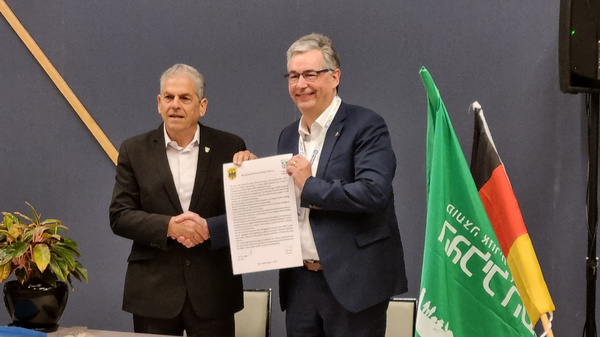 Deux hommes se serrent la main, l'un tenant un document en papier. Ils sont devant une table. En arrière-plan, des drapeaux et une plante décorative. L'ambiance semble formelle, suggérant un accord ou une coopération.