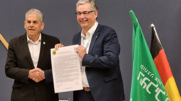Zwei Herren stehen an einem Tisch und schütteln sich die Hände. Der Mann auf der linken Seite hält eine Urkunde in der Hand. Im Hintergrund sind eine grüne und eine deutsche Flagge sowie eine Pflanze zu sehen.