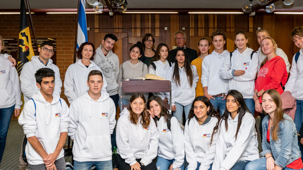 A diverse group of about 25 young adults is gathered in a room, wearing matching hoodies. They are posing together around a table, with some flags in the background. The atmosphere appears friendly and celebratory, indicating a group event or gathering.