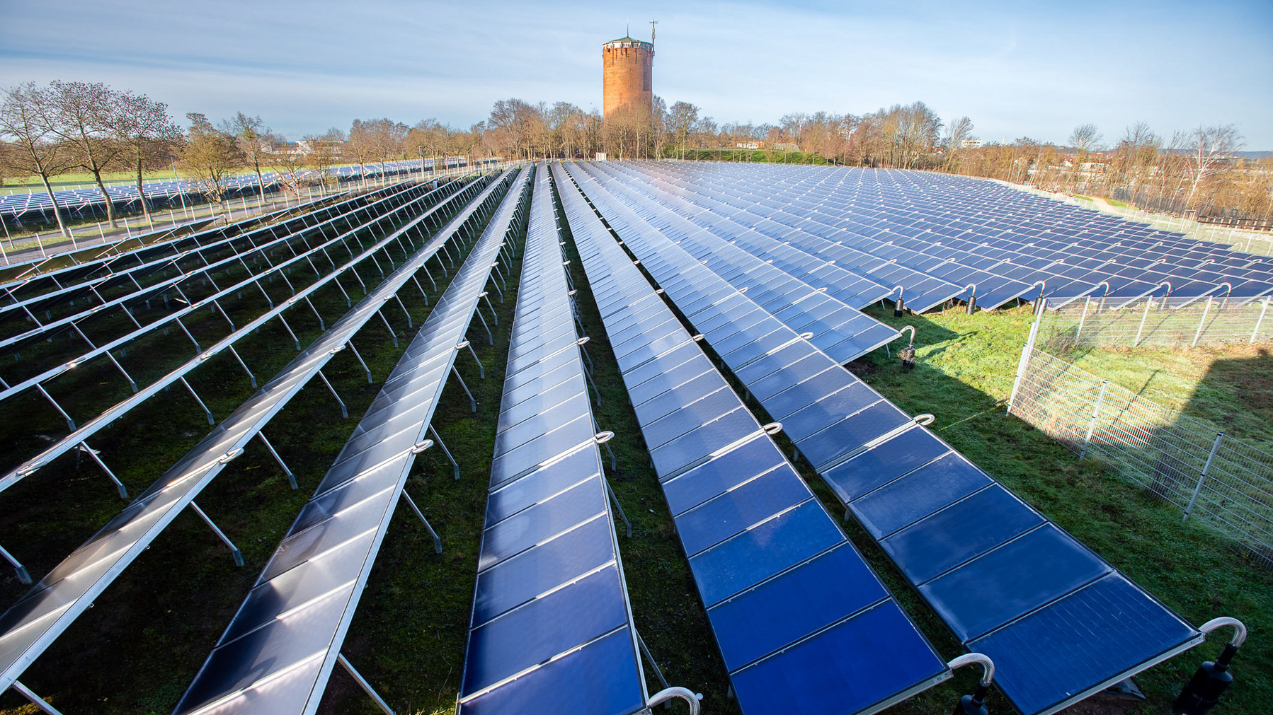 Ein Solarpark mit vielen Photovoltaikanlagen, die in Reihen aufgestellt sind, erstreckt sich über eine grüne Wiese. Im Hintergrund steht ein alter Wasserturm. Die Aufnahme zeigt den Fokus auf der nachhaltigen Energieerzeugung durch Solarpanel.