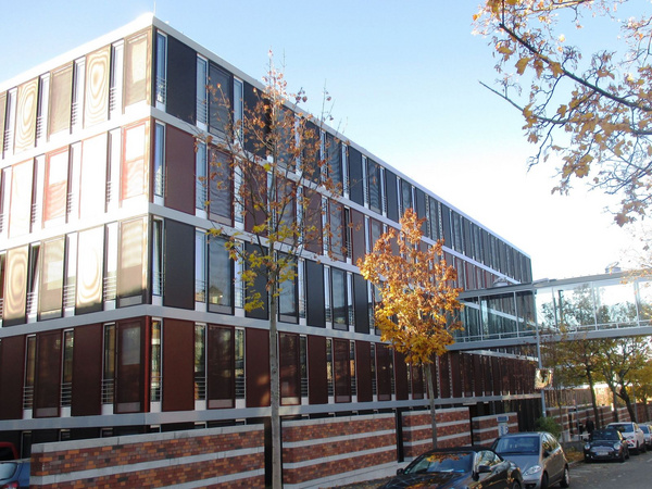 Ein modernes, mehrstöckiges Gebäude mit einer Fassade aus roten und weißen Elementen. Große Fensterfronten sorgen für viel Licht. Vorn sind einige Bäume zu sehen, deren Blätter orange und gelb gefärbt sind, was auf den Herbst hinweist. Ein Glasgang verbindet das Gebäude mit einem weiteren.