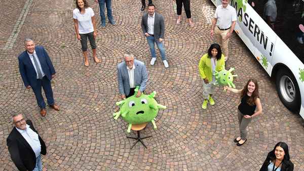 A diverse group of people stands on a cobblestone plaza, some in business attire and others in casual clothing. A large, friendly virus mascot is featured prominently in the foreground, symbolizing a health awareness campaign focused on vaccinations, with a bus in the background advertising "Stop Measles!"