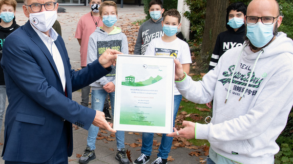 A group of students wearing face masks stands together outside a building, while two adults, also masked, hold a certificate. The setting conveys a sense of achievement, likely related to a school or community project.