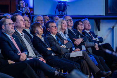 Eine Gruppe von Menschen sitzt in einem Publikum und hört aufmerksam zu. Die Anwesenden sind in formeller Kleidung und zeigen unterschiedliche Gesichtsausdrücke, die von Interesse bis zur Nachdenklichkeit reichen. Die Atmosphäre wirkt gespannt und engagiert.