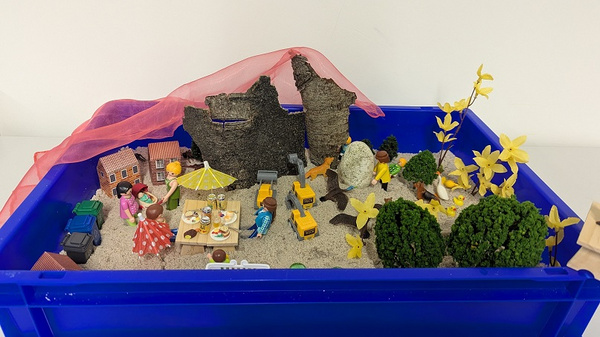 Eine blaue Kiste mit Sand enthält Szenen aus dem Alltag. Figuren stehen um einen Tisch mit Essen, während andere an einem Baustellenfahrzeug arbeiten. Kleine Bäume und gelbe Blumen sind verteilt. Im Hintergrund sind zerfallene Mauern und ein durchscheinender, roter Stoff sichtbar.