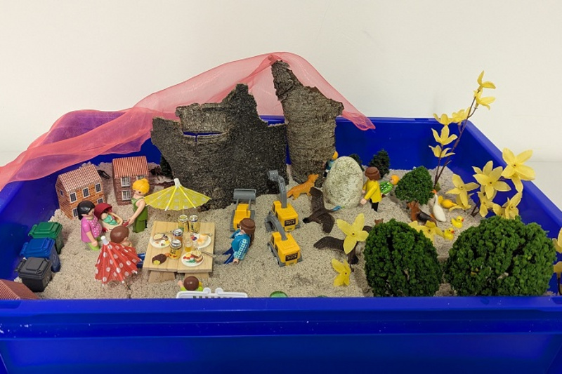 Eine blaue Kiste mit Sand enthält Szenen aus dem Alltag. Figuren stehen um einen Tisch mit Essen, während andere an einem Baustellenfahrzeug arbeiten. Kleine Bäume und gelbe Blumen sind verteilt. Im Hintergrund sind zerfallene Mauern und ein durchscheinender, roter Stoff sichtbar.