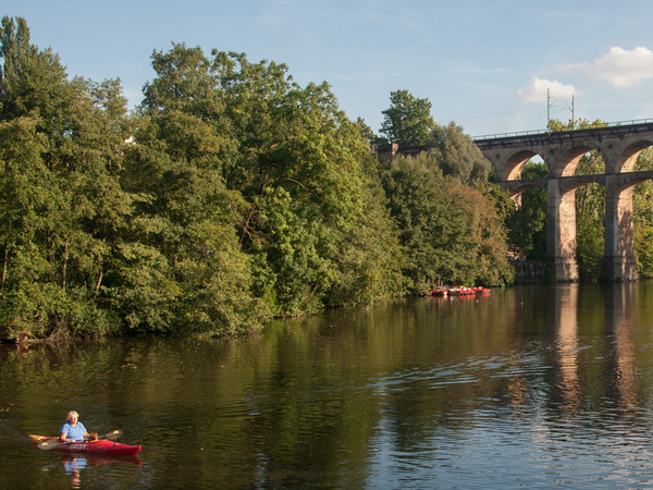Ein Kajakfahrer paddelt auf einem ruhigen Gewässer, umgeben von dichtem Grün. Im Hintergrund erhebt sich ein imposantes Viadukt mit mehreren Bögen, das über den Fluss führt. Der Himmel ist klar mit wenigen Wolken.