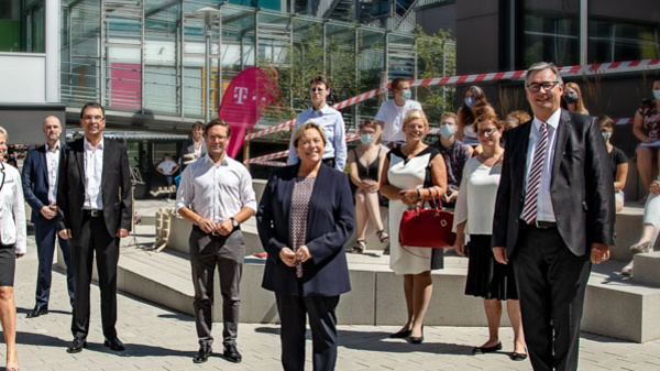 A group of professionals stands on a paved outdoor area of a modern building, smiling and posing for the photo. They are dressed in formal attire, with some holding papers. In the background, there are people visible on an upper level, and a colorful sign can be seen.