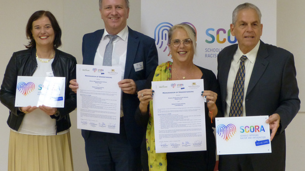 Quatre personnes se tiennent debout, tenant des certificats. Elles sourient, debout devant un panneau avec le logo de SCORA. L'image évoque une cérémonie officielle, axée sur la sensibilisation à la discrimination raciale.