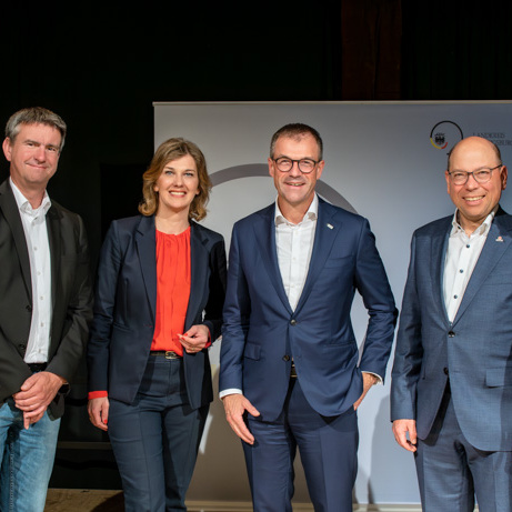 Sechs Personen stehen in einer Reihe, lächeln und posieren für ein Gruppenfoto. Sie tragen formelle Kleidung und stehen vor einem weißen Hintergrund mit einem Logo. Die Szene vermittelt eine positive, professionelle Atmosphäre.