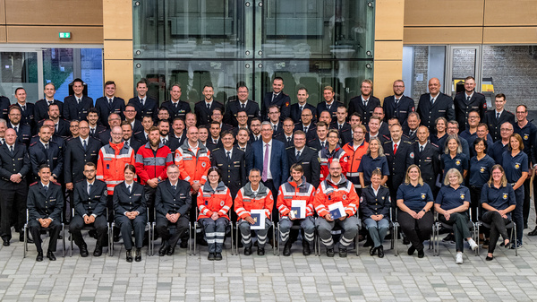 Gruppenfoto von etwa 75 Personen in verschiedenen Uniformen, darunter Feuerwehr, Rettungsdienst und Zivilkleidung. Sie stehen vor einem modernen Gebäude. Die Atmosphäre ist freundlich und professionell, mit dem Fokus auf Teamarbeit und Gemeinschaft.