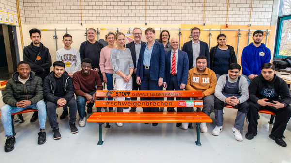 Eine Gruppe von jungen Menschen und Erwachsenen steht und sitzt in einem Raum, umgeben von Bänken. Sie posieren vor einer orangefarbenen Bank mit dem Text „STopp Gewalt gegen Frauen - #schweigenbrechen“, um ihr Engagement gegen Gewalt auszudrücken.