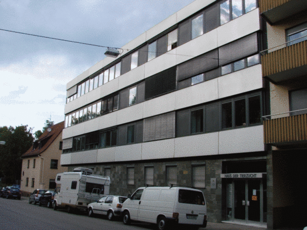 Ein modernes Gebäude mit mehreren Stockwerken und großen Fenstern, das an einer Straße steht. Die Fassade ist überwiegend in Weiß und Grau gehalten. Vor dem Gebäude parken mehrere Fahrzeuge, darunter ein Wohnmobil und weiße Lieferwagen.