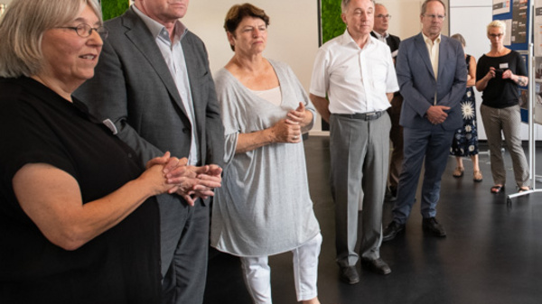 A group of six people is engaged in conversation near display boards in a modern indoor setting. The individuals, dressed in business attire, appear to be discussing the information presented on the boards, which feature various documents and images.