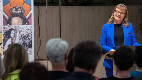 Eine Frau in einem blauen Outfit steht am Redepult und spricht zu einem Publikum. Im Hintergrund ist ein Banner mit Kunstmotiven und dem Text "KUNST im Kreishaus" zu sehen. Die Zuhörer sind teils von hinten sichtbar und scheinen dem Vortrag aufmerksam zuzuhören.